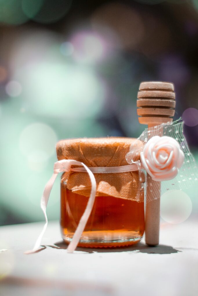 A clear glass jar filled with golden honey, adorned with ribbons and a wooden dipper, evoking sweetness and elegance.